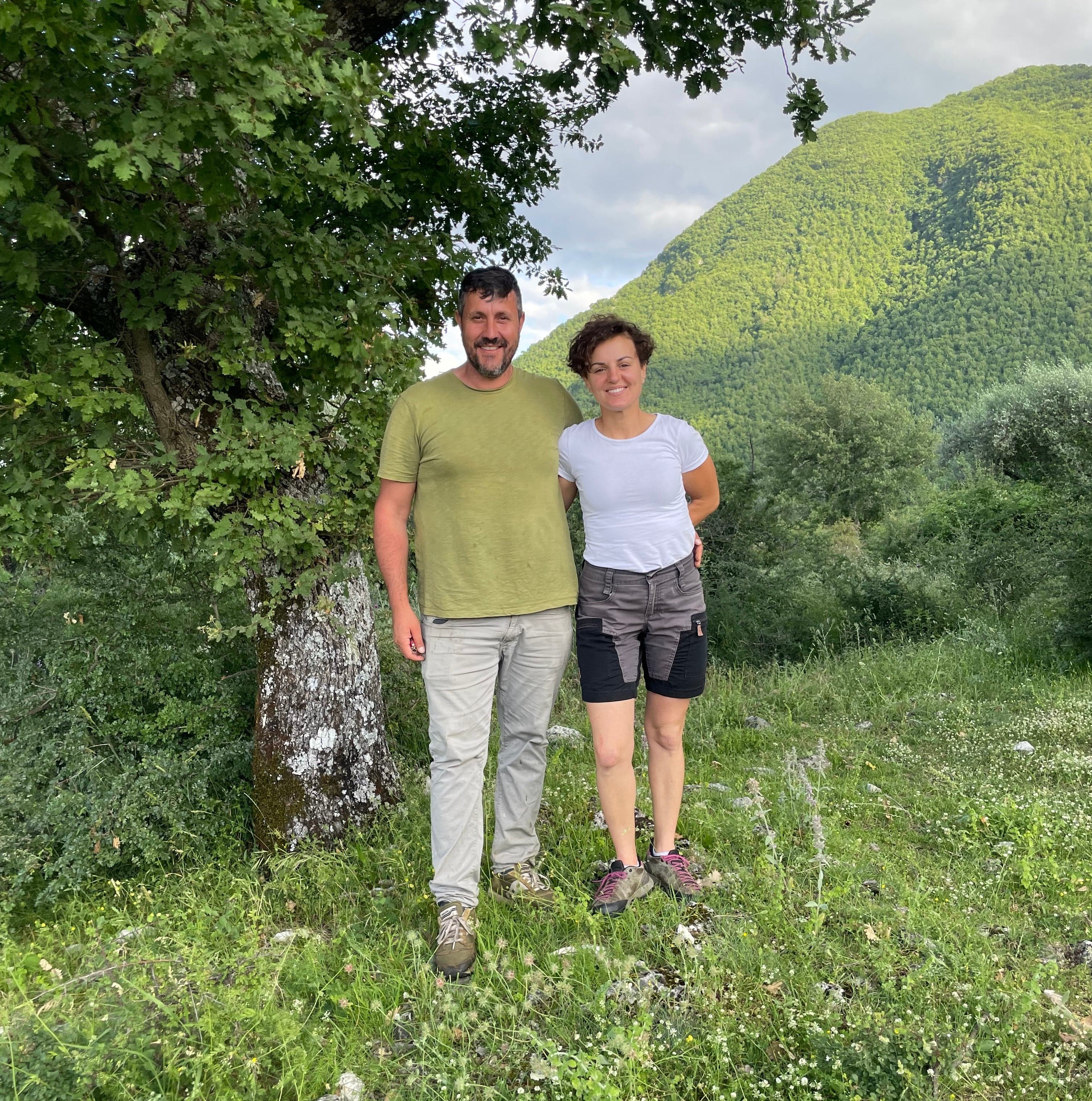 Two people standing in a grassy field with green hills and trees in the background