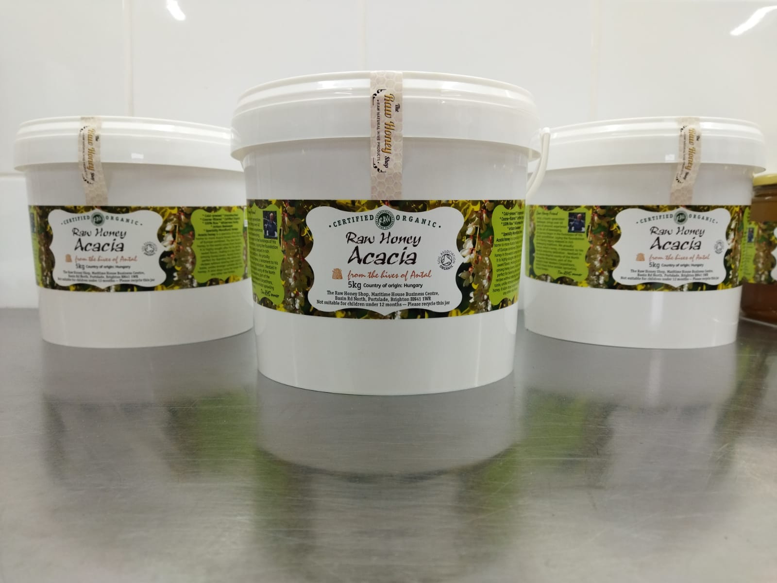 Three white buckets with green and yellow labels, indicating raw certified organic acacia honey.