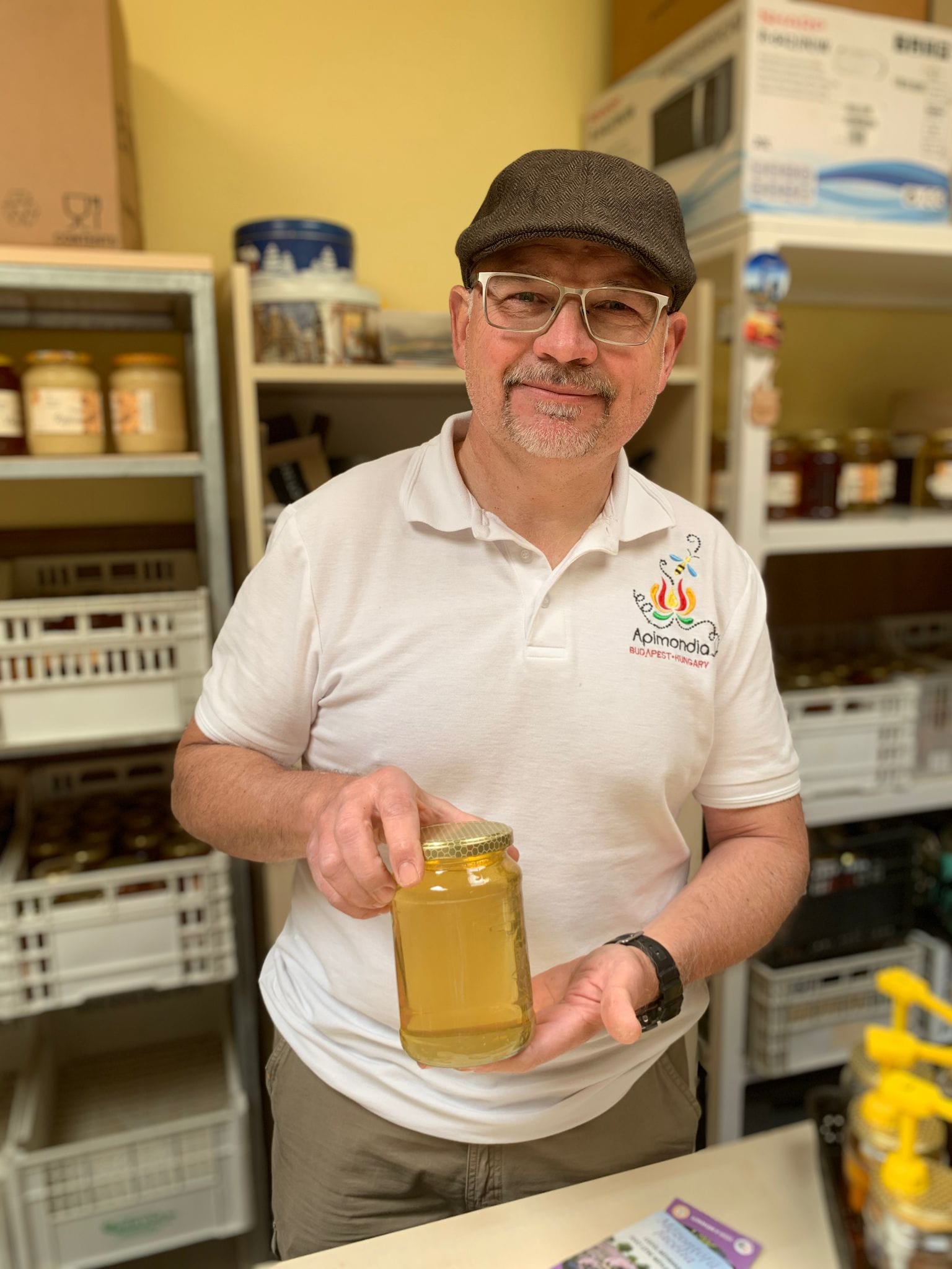 Beekeeper Antal wearing a flat cap and glasses.  Wearing a white t-shirt and holding a jar of his honey.