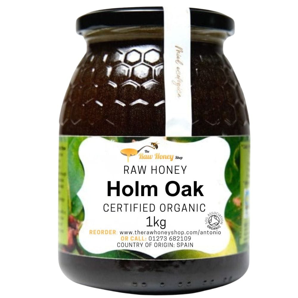 A 1kg glass jar of The Raw Honey Shop Holm Oak Raw Honey, featuring a dark honey-colored product inside, a black lid, and a green and white label.