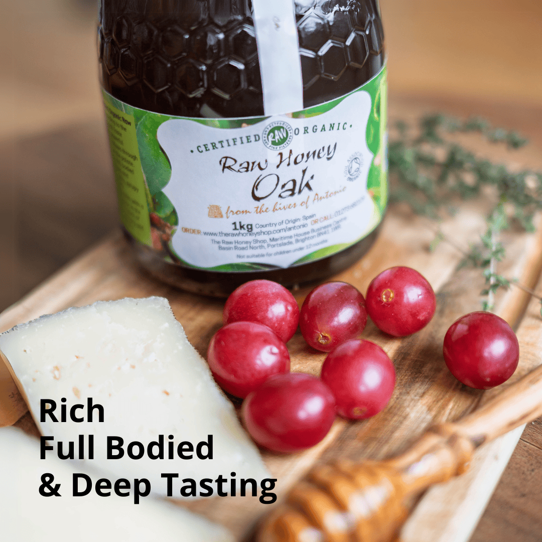 A jar of raw honey oak honey from Certified Raw Organic, styled on a wooden board with a slice of cheese, red grapes, and herbs. Text overlay reads 'Rich Full Bodied & Deep Tasting'.