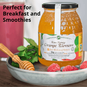 A jar of Organic Raw Honey Orange Blossom honey displayed on a wooden table with a metal bowl containing raspberries, blueberries, mint leaves, a honey dipper, and a blurred pink smoothie in the background. Text overlay reads 'Perfect for Breakfast and Smoothies'.