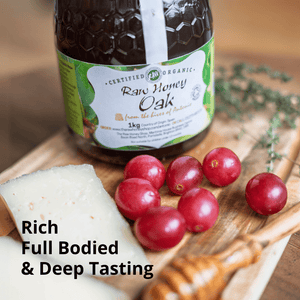 A jar of raw organic raw honey oak honey displayed on a wooden board with a slice of white cheese and red grapes. Text overlay reads 'Rich Full Bodied & Deep Tasting'.