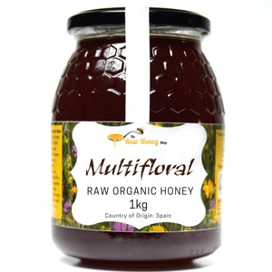 A 1kg glass jar of Antonios Multifloral Raw Organic Honey from The Raw Honey Shop. On a white background, with a vertical white stripe running down the center of the glass.