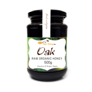 A 500g glass jar of Antonios Oak Raw Organic Honey from The Raw Honey Shop, featuring a black lid, a white vertical label, and a green and white decorative front label.