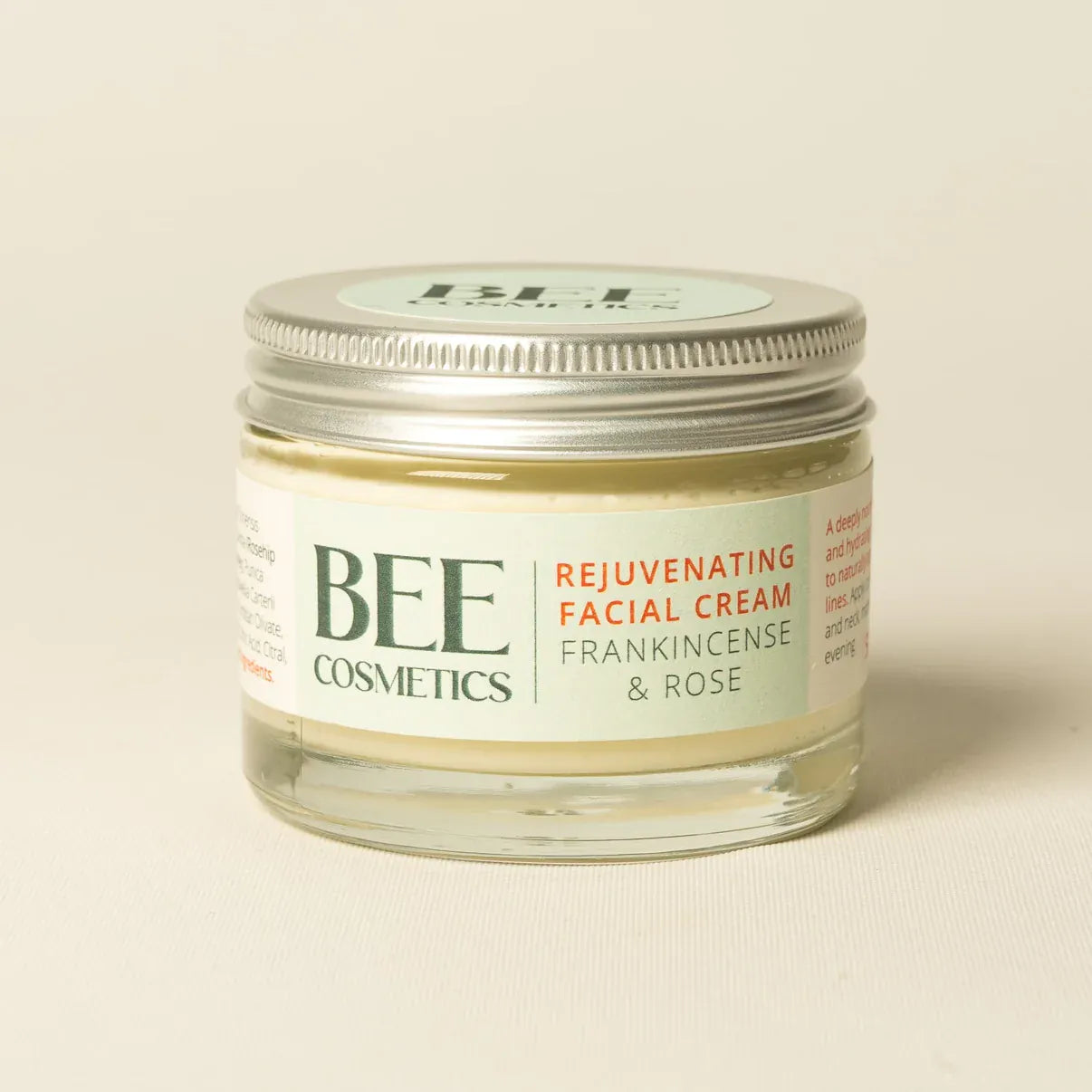 A small clear glass jar of BEE COSMETICS Rejuvenating Facial Cream with Frankincense & Rose, featuring a silver metal lid and a cream colored label with green and orange text, set against a plain light beige background.