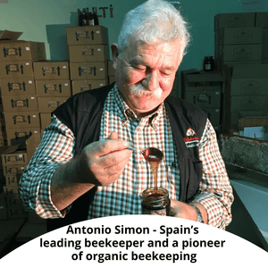 Antonio Simon, Spain's leading beekeeper, shown holding a jar of honey and lifting a spoonful of the golden liquid. He is wearing a black vest over a red and white checkered shirt, with stacks of cardboard boxes in the background.