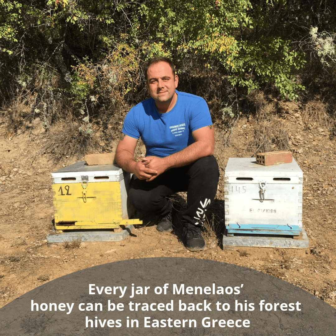 A man in a blue t-shirt and black pants kneels outdoors between two wooden beehives, one yellow and one white, with trees and dry grass in the background. A text overlay at the bottom reads, 'Every jar of Menelaos' honey can be traced back to his forest hives in Eastern Greece'.