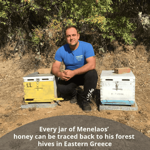 A man in a blue t-shirt and black pants kneels outdoors between two wooden beehives, one yellow and one white, with trees and dry grass in the background. A text overlay at the bottom reads, 'Every jar of Menelaos' honey can be traced back to his forest hives in Eastern Greece'.
