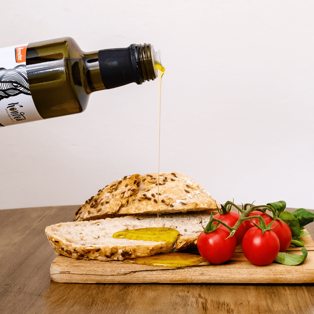Bottle of olive oil being poured over bread and tomatoes on a wooden board.