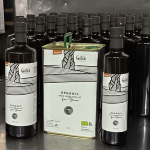 Tin of olive oil with white label surrounded by dark bottles of olive oil