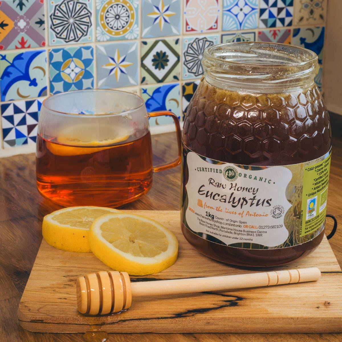 A jar of raw organic eucalyptus honey sits on a wooden cutting board next to a glass mug of tea with a lemon slice and two loose lemon slices. A wooden honey dipper rests in front of the jar, with a colorful tiled backsplash behind the scene.