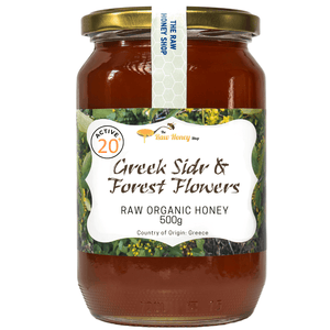 A 500g glass jar of Active 20 Greek Sidr & Forest Flowers Raw Organic Honey by The Raw Honey Shop, featuring a gold metal lid and a white label with green and yellow accents, set against a plain white background.