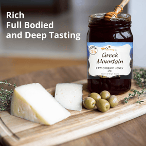 A jar of Active 15 Greek Mountain Raw Organic Honey displayed on a wooden cutting board with cheese wedges, olives, and herbs, alongside text overlay reading 'Rich Full Bodied and Deep Tasting'.