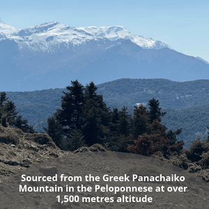 Mountain landscape with snow-capped peaks, blue haze in the distance, dark evergreen trees in the foreground, and a dirt path at the bottom. White text at the bottom reads: 'Sourced from the Greek Panachaiko Mountain in the Peloponnese at over 1,500 metres altitude'.