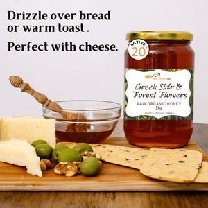 A jar of Greek Sidr & Forest Flowers raw organic honey stands on a wooden board with cheese, olives, crackers, and a small glass bowl of honey with a wooden dipper. Text at the top reads, 'Drizzle over bread or warm toast.