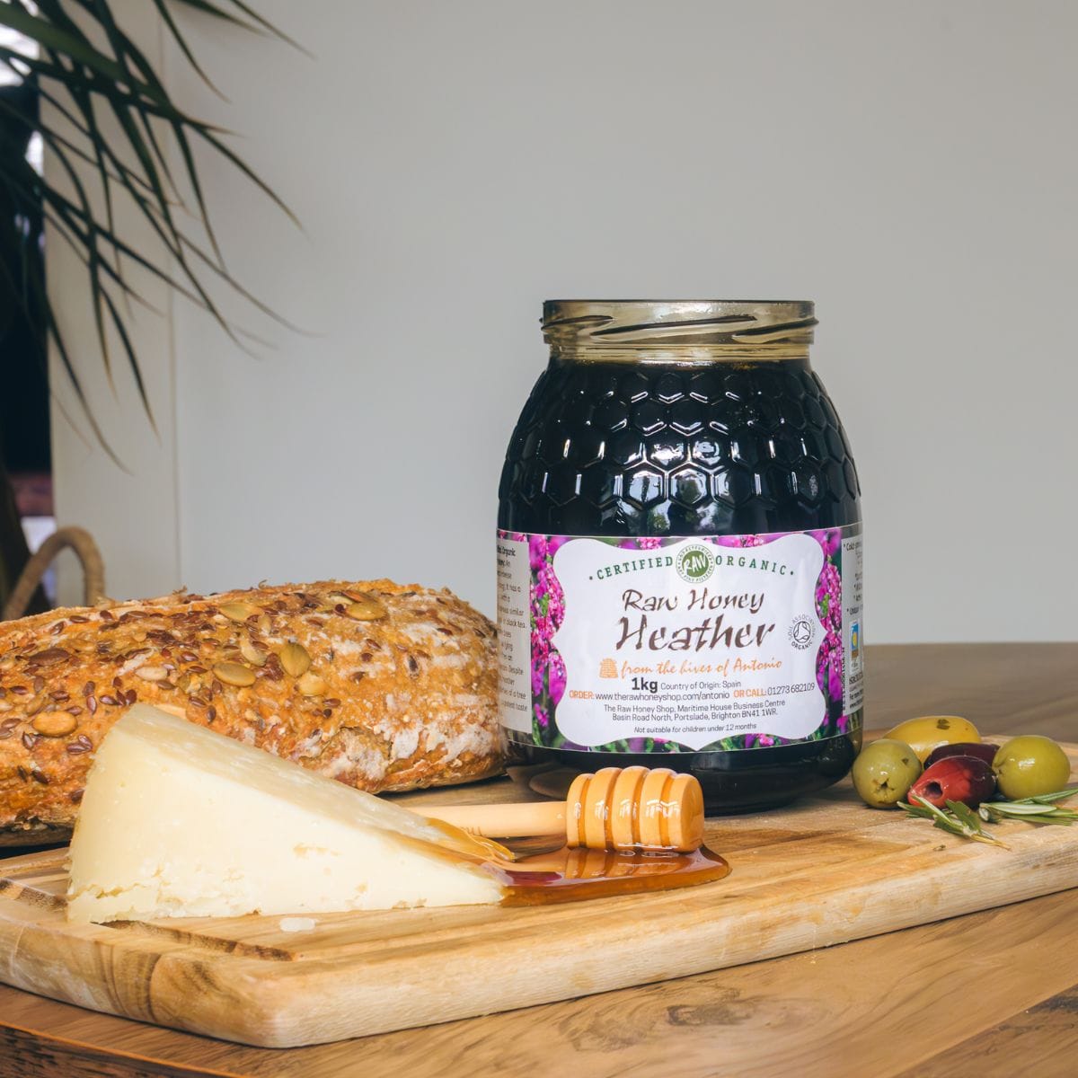 A jar of Certified Organic Raw Honey Heather on a wooden cutting board with a slice of bread, a wedge of cheese, honey, and olives, set against a neutral indoor background.