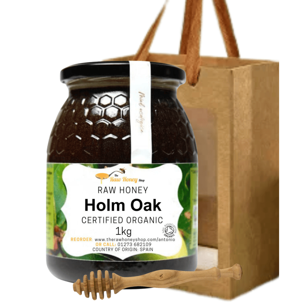 A jar of raw organic Holm Oak honey from Taste Honey Shop, shown in front of a brown paper gift bag with wooden handles and a wooden honey dipper, on a plain white background.