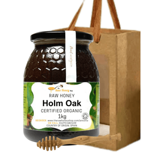 A jar of raw organic Holm Oak honey from Taste Honey Shop, shown in front of a brown paper gift bag with wooden handles and a wooden honey dipper, on a plain white background.