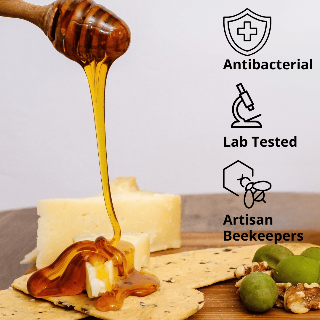 A close-up of amber honey dripping from a wooden honey dipper onto cheese and crackers on a wooden board, with text callouts reading Antibacterial, Lab Tested, and Artisan Beekeepers.