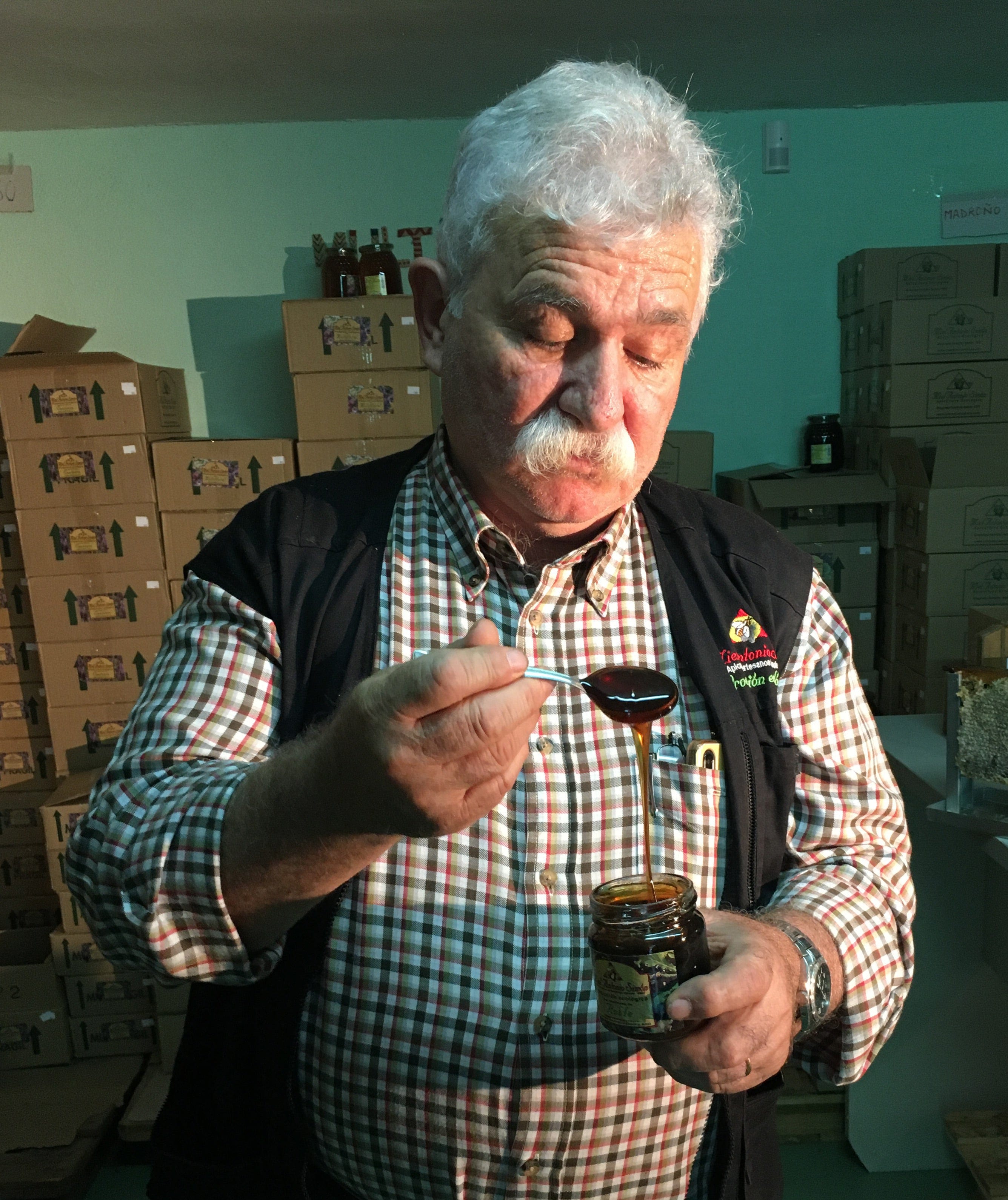 Man with silver hair holding a jar of honey and a spoon with honey dripping from the spoon.