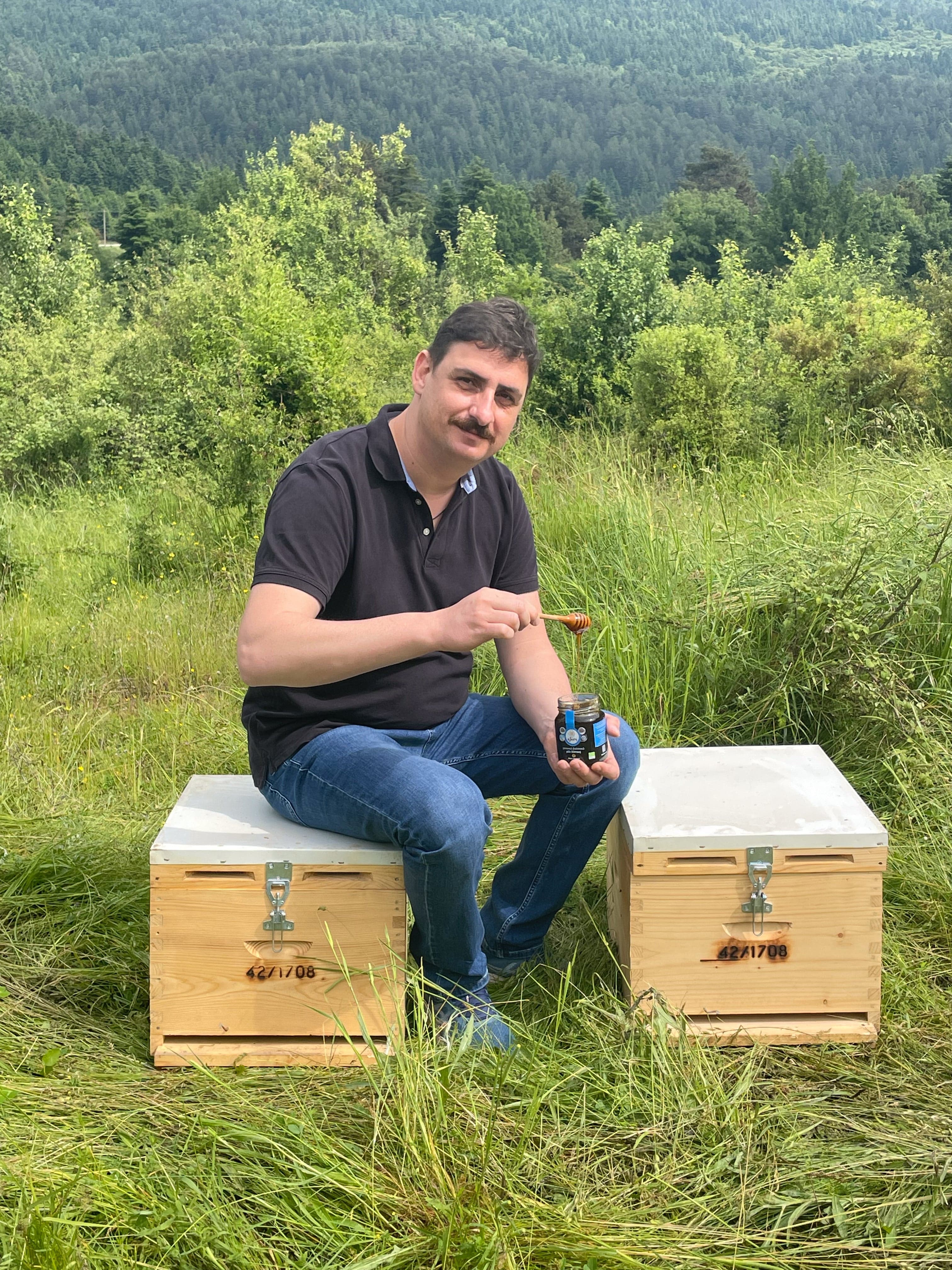 Beekeeper Dimitris sitting on hive holding jar of Mount Olympus mountain honey.