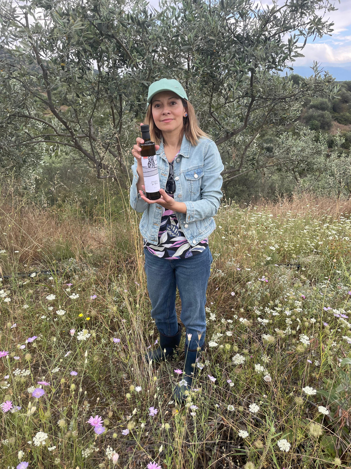 Image of lady in a field of wild flowers with an olive tree in the background holding a bottle of olive oil.