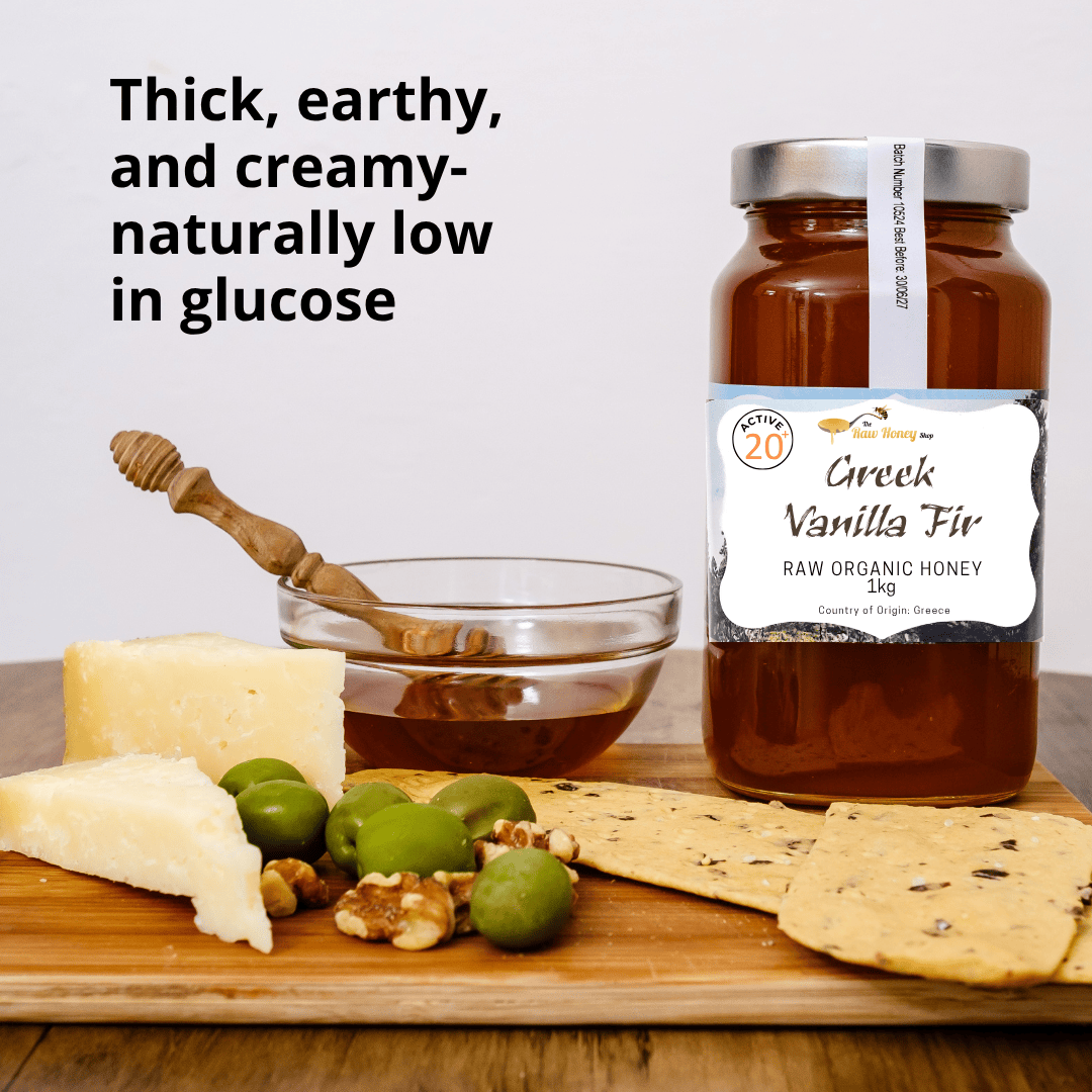 A jar of Greek Vanilla Fir raw organic honey by Raw Honey Shop is displayed on a wooden board with cheese, olives, walnuts, and crackers, alongside a glass bowl of honey and a wooden honey dipper. Text on the left reads 'Thick, earthy, and creamy-naturally low in glucose'.