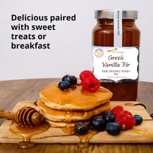 A jar of Greek Vanilla Fir raw organic honey displayed behind a stack of pancakes topped with blueberries and raspberries on a wooden board, with a honey dipper and syrup drizzle. Text on the left reads 'Delicious paired with sweet treats or breakfast'.