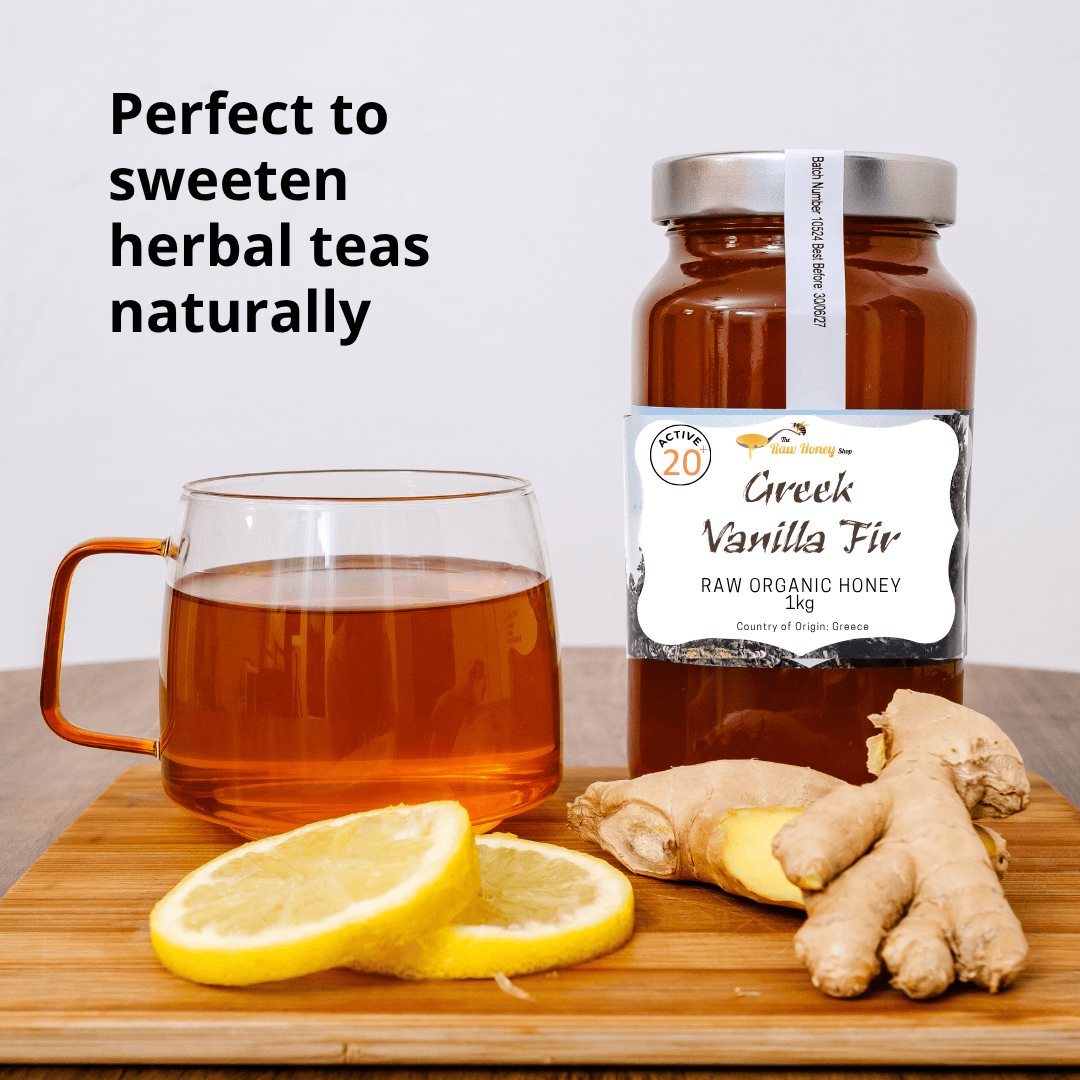 A jar of Greek Vanilla Fir raw organic honey by Honey by the Sea sits on a wooden cutting board next to a glass mug filled with amber herbal tea, sliced lemons, and ginger roots. Text on the left reads 'Perfect to sweeten herbal teas naturally'.