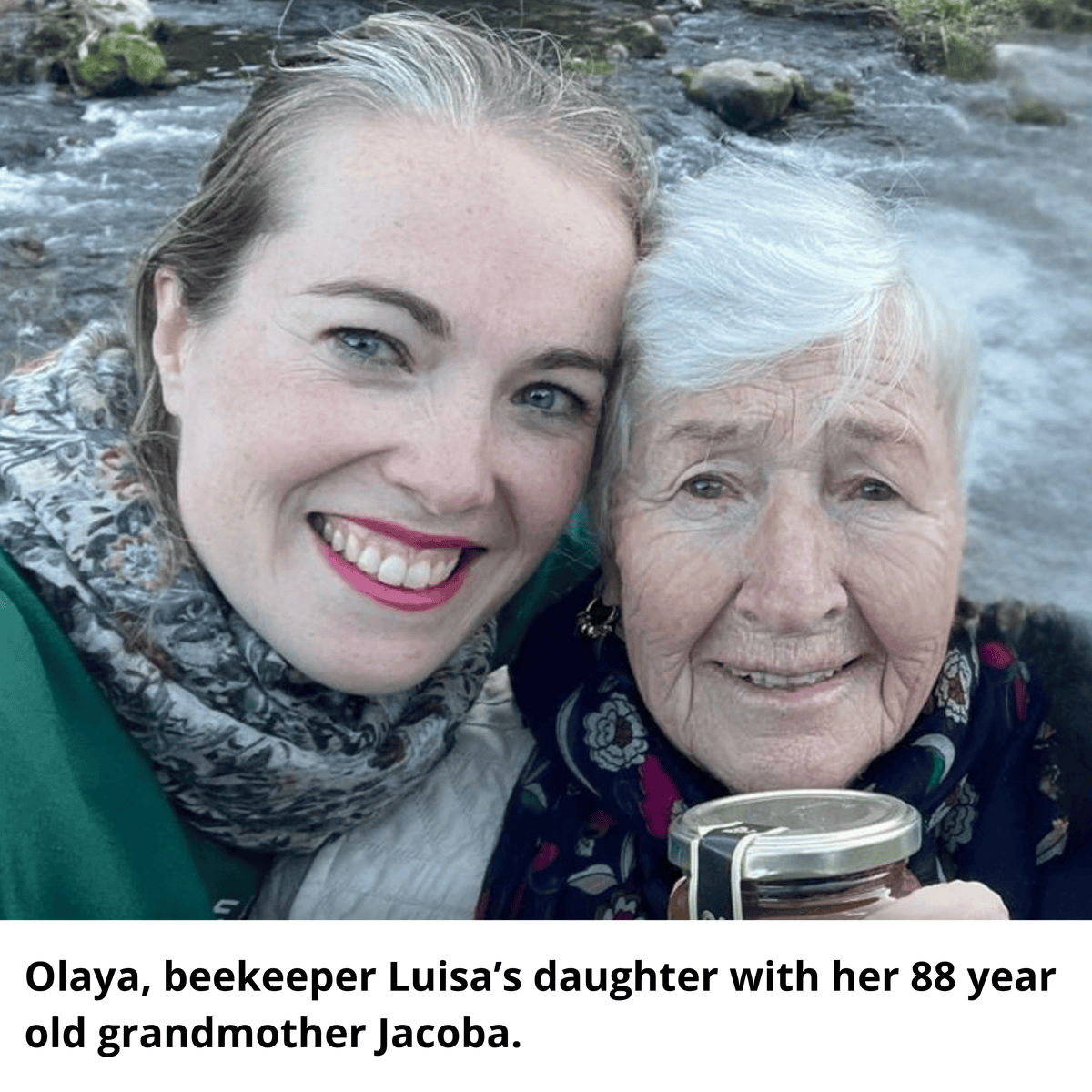 A close-up photo of two women smiling outdoors near a flowing stream, with a text overlay at the bottom identifying them as Olaya, beekeeper Luisa's daughter, and her 88-year-old grandmother Jacoba.