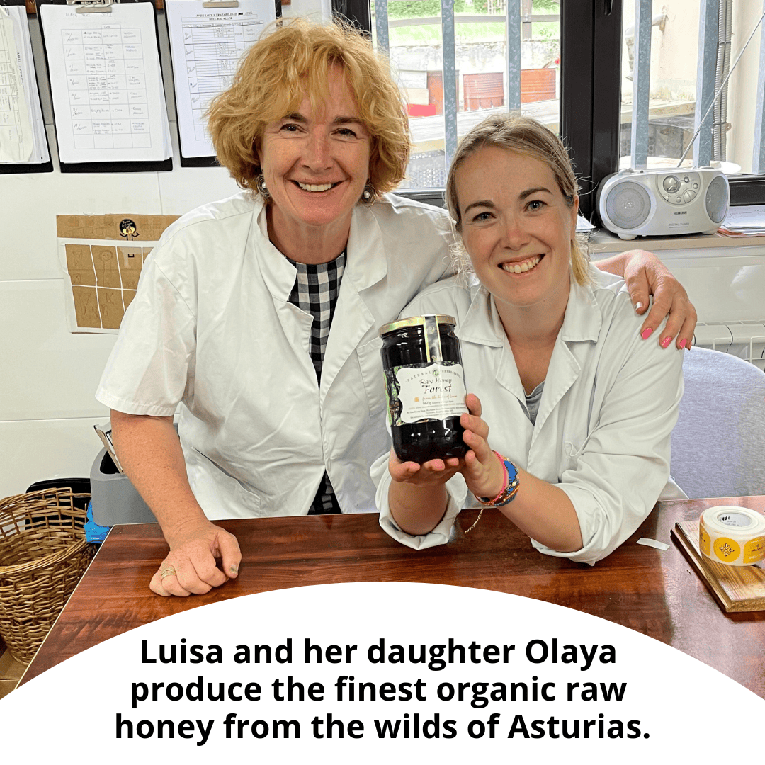 Two women in white lab coats smile while holding a jar of raw honey on a wooden table. A text overlay at the bottom reads, 'Luisa and her daughter Olaya produce the finest organic raw honey from the wilds of Asturias.