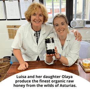 Two women in white lab coats smile while holding a jar of raw honey on a wooden table. A text overlay at the bottom reads, 'Luisa and her daughter Olaya produce the finest organic raw honey from the wilds of Asturias.