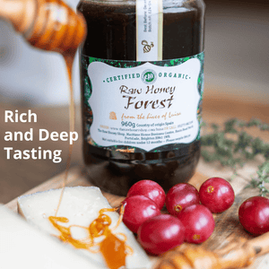 A bottle of Certified Organic Raw Honey Forest honey sits on a wooden board with a honey dipper pouring honey over cheese and red cranberries. Text on the left reads 'Rich and Deep Tasting'.