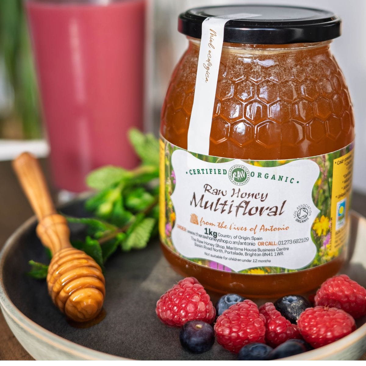 A jar of certified organic raw honey multifloral from the trees of Antonio, displayed on a dark plate with fresh raspberries, blueberries, a wooden honey dipper, and mint leaves.