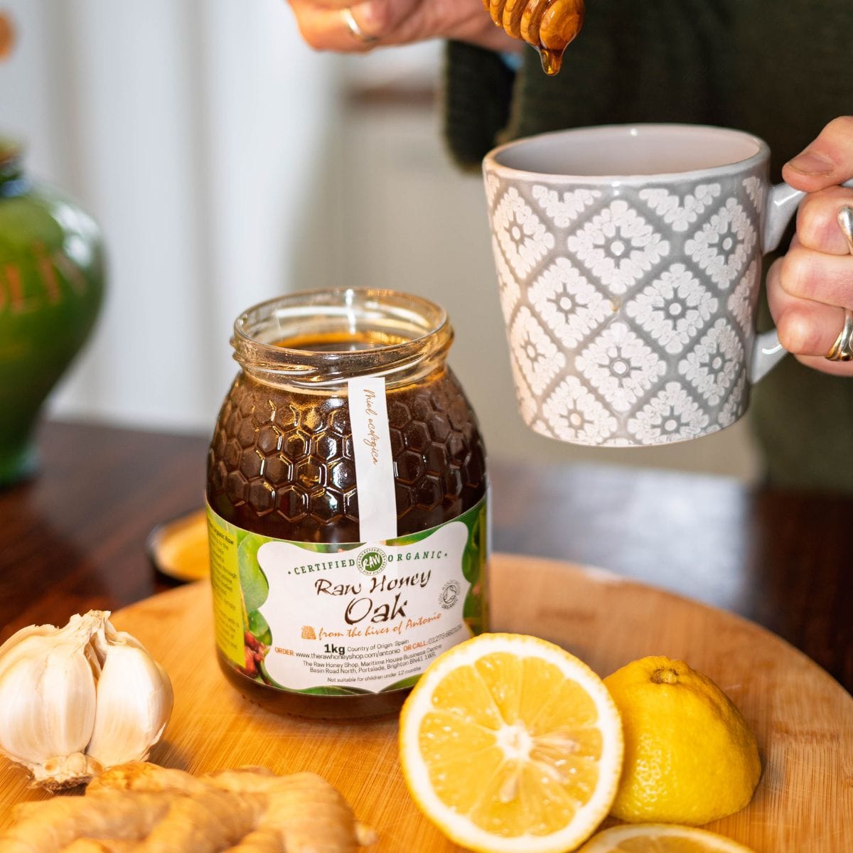 A jar of Centfused Organics Raw Honey Oak raw unfiltered honey sits on a wooden cutting board with lemons, ginger, and garlic, while a person holds a patterned mug above the scene.