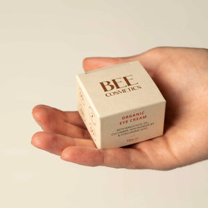 A hand holding a small beige square box of BEE COSMETICS Organic Eye Cream against a plain light beige background.