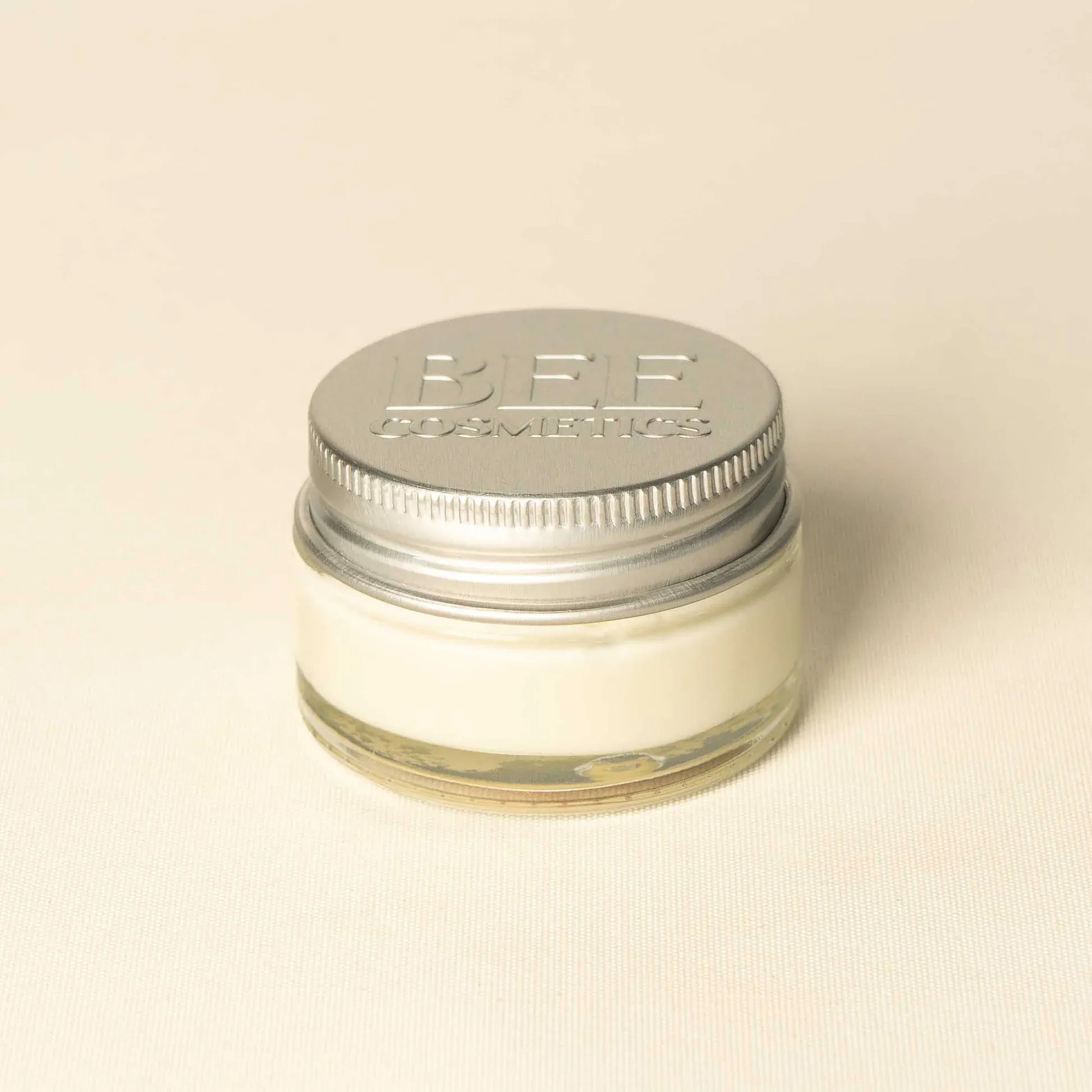 A small clear glass jar filled with off-white cream or balm, sealed with a silver metal screw lid embossed with 'BEE COSMETICS', on a plain light beige background.