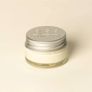 A small clear glass jar filled with off-white cream or balm, sealed with a silver metal screw lid embossed with 'BEE COSMETICS', on a plain light beige background.