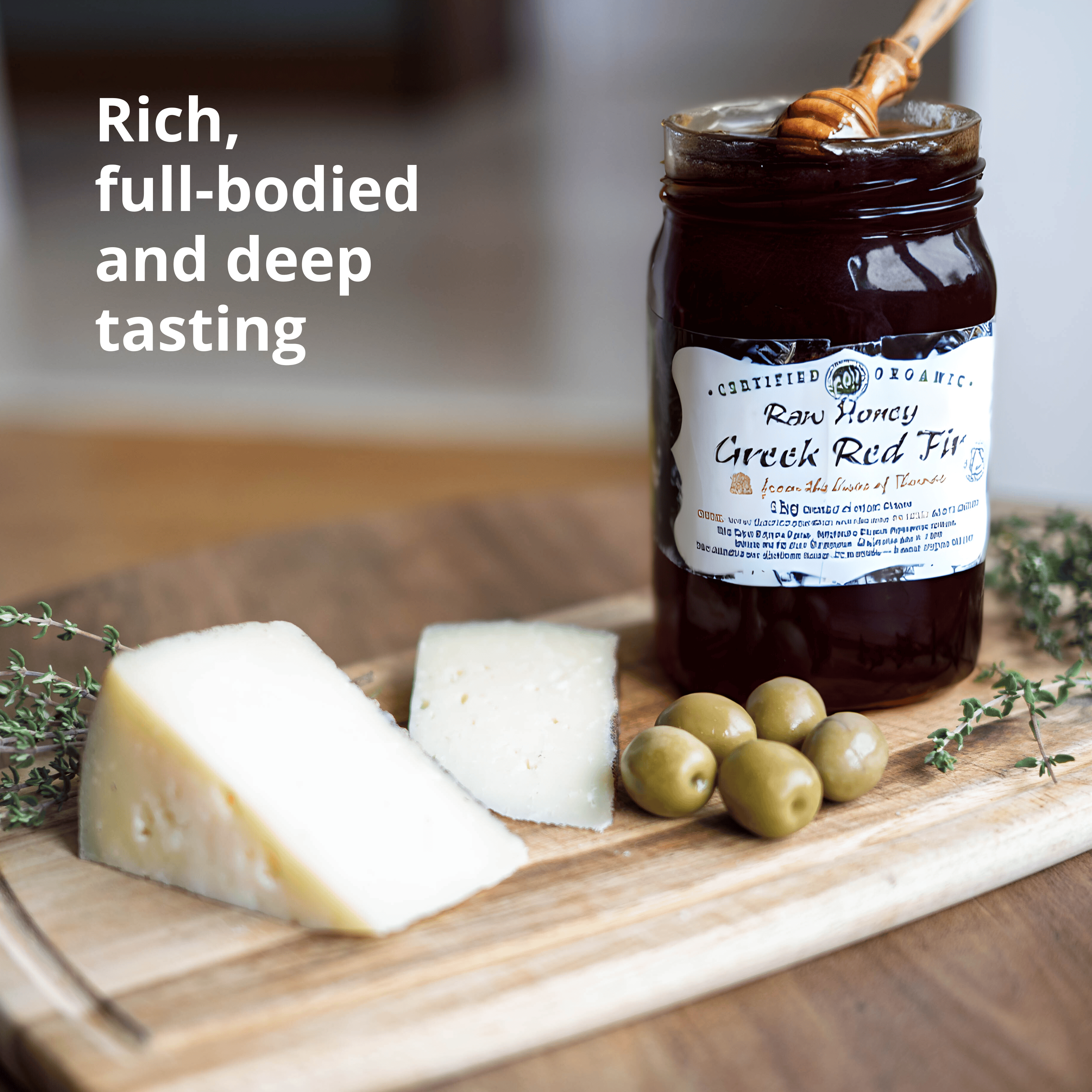 A jar of Organic Raw Honey Greek Rid Fir honey displayed on a wooden cutting board with cheese wedges, green olives, and sprigs of thyme. The background is a warm, softly blurred kitchen setting, and the text 'Rich, full-bodied and deep tasting' is visible on the left.