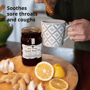 A jar of Organic Raw Honey Greek Red Top honey sits on a wooden cutting board with ginger, garlic, and lemon slices, next to a person holding a patterned mug and a honey dipper. Text on the image reads 'Soothes sore throats and coughs'.