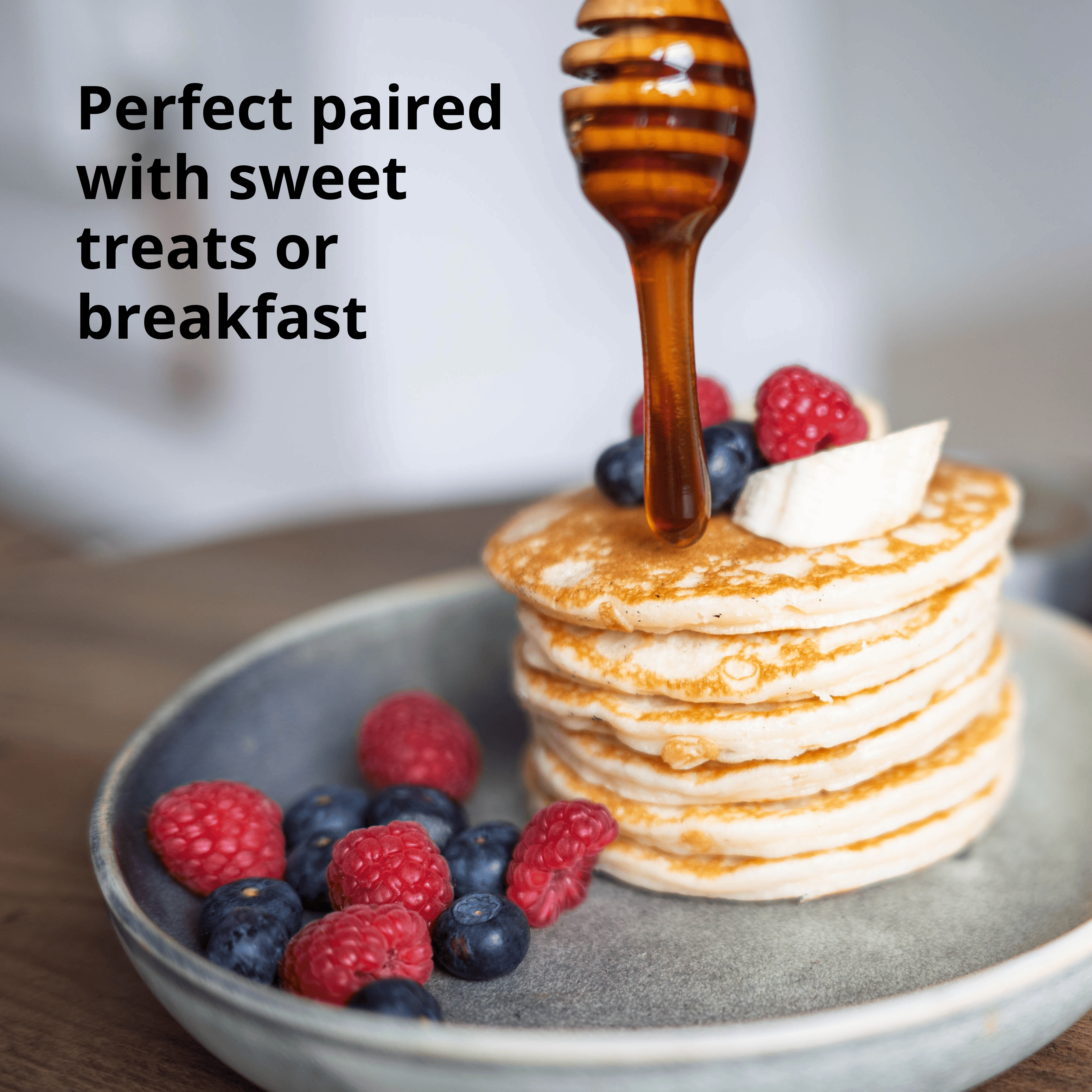 A stack of pancakes topped with a dollop of cream and fresh berries, with honey dripping from a wooden dipper. The plate sits on a wooden surface, and the text reads 'Perfect paired with sweet treats or breakfast'.