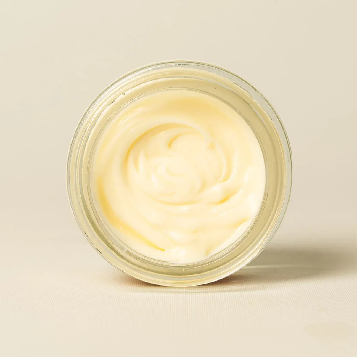 An open clear glass jar filled with a pale creamy yellow whipped or melted product, photographed from above on a plain warm off-white background.
