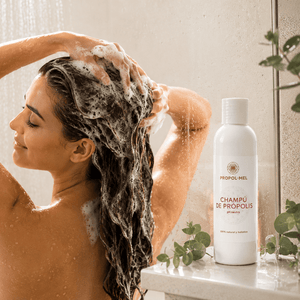 A woman washing her hair in a shower with lathered hair, beside a white bottle labeled PROPOL-MEL Champü de Propólis on a bathroom ledge with greenery.