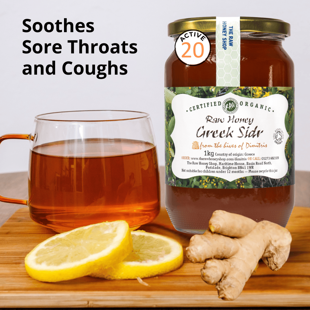 A jar of Raw Honey Creek Slid organic honey stands on a wooden surface beside a glass mug filled with amber tea, lemon slices, and ginger roots. Text on the left reads 'Soothes Sore Throats and Coughs.