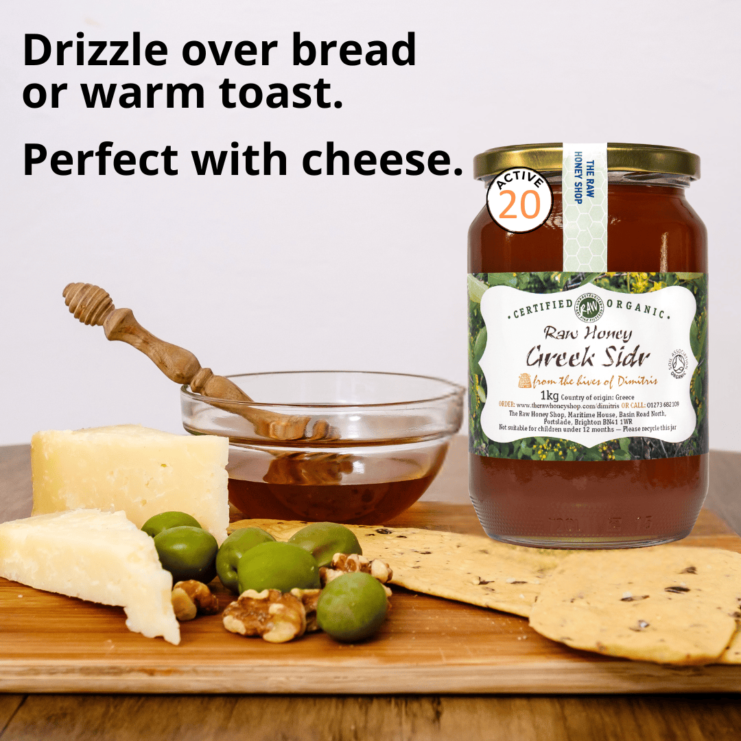 A jar of Certified Organic Raw Honey Greek Sider honey displayed on a wooden board with cheese, olives, walnuts, and flatbread, alongside a honey dipper and a glass bowl of honey. Text on the image reads 'Drizzle over bread or warm toast.