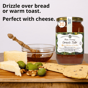 A jar of Certified Organic Raw Honey Greek Sider honey displayed on a wooden board with cheese, olives, walnuts, and flatbread, alongside a honey dipper and a glass bowl of honey. Text on the image reads 'Drizzle over bread or warm toast.