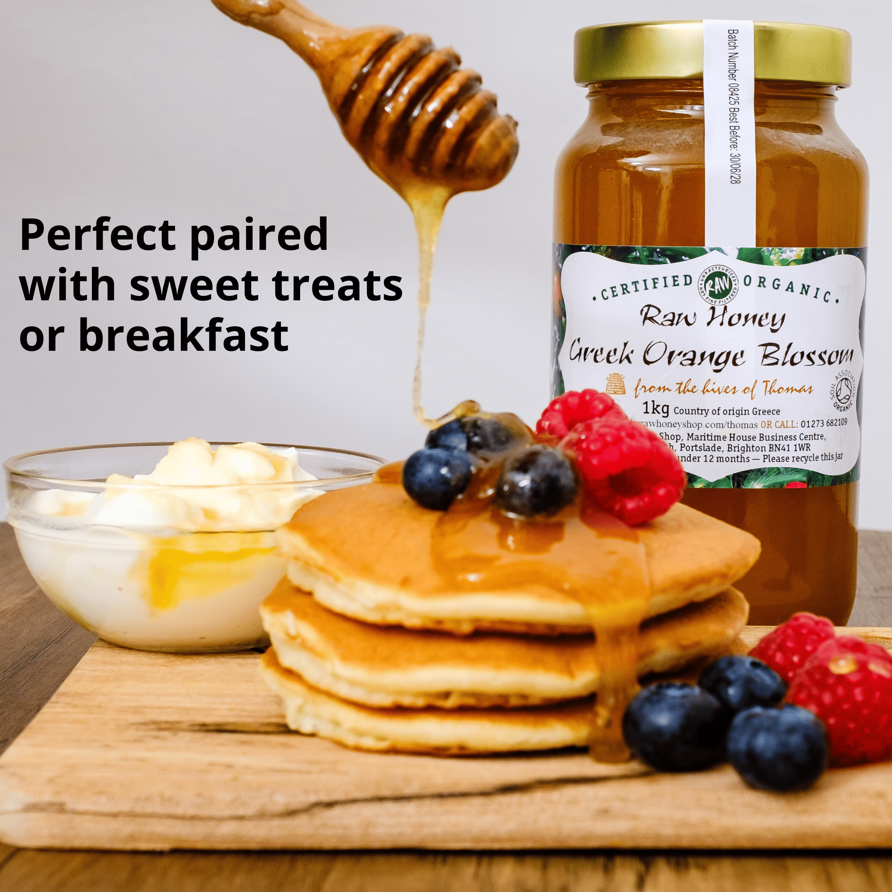A jar of raw honey Greek orange blossom jam stands on a wooden board beside pancakes topped with syrup and berries, a bowl of butter, and a honey dipper, with text reading 'Perfect paired with sweet treats or breakfast.'.