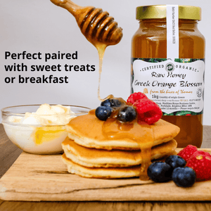 A jar of raw honey Greek orange blossom jam stands on a wooden board beside pancakes topped with syrup and berries, a bowl of butter, and a honey dipper, with text reading 'Perfect paired with sweet treats or breakfast.'.