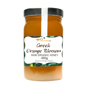 A jar of Greek Orange Blossom Raw Organic Honey from The Raw Honey Shop, featuring a black lid and a white label with green accents, set against a plain white background.